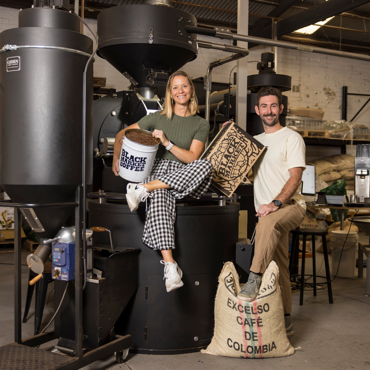 Two people sitting on coffee roasting equipment with a 'Black Market Coffee' box 